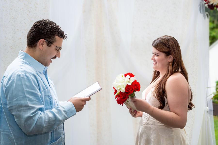 A groom reads his vows to his bride, who holds a bouquet of red and white roses